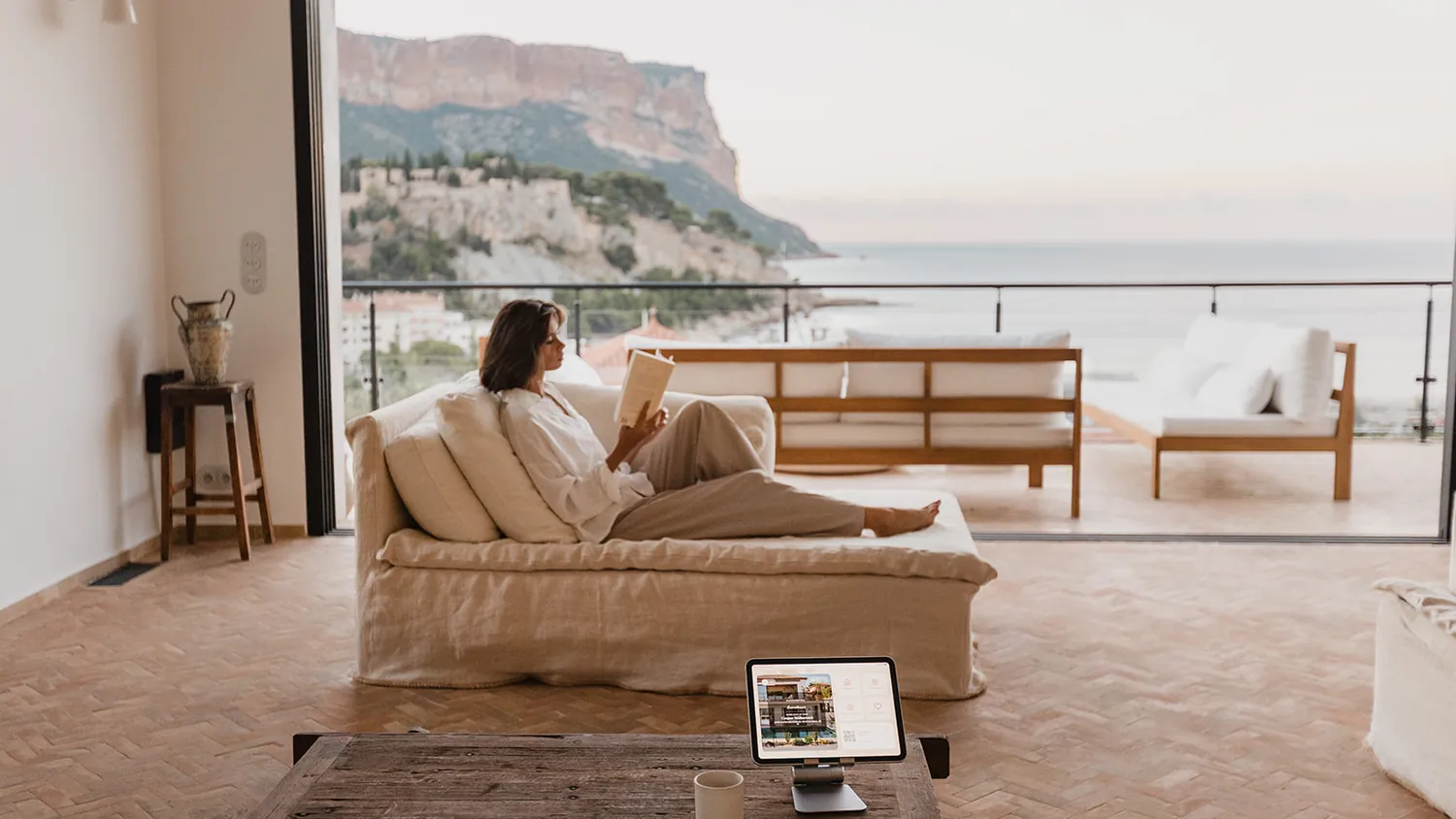Une voyageuse lit sur le canapé, face aux falaises de Cassis, le guide ByOhana posé sur la table